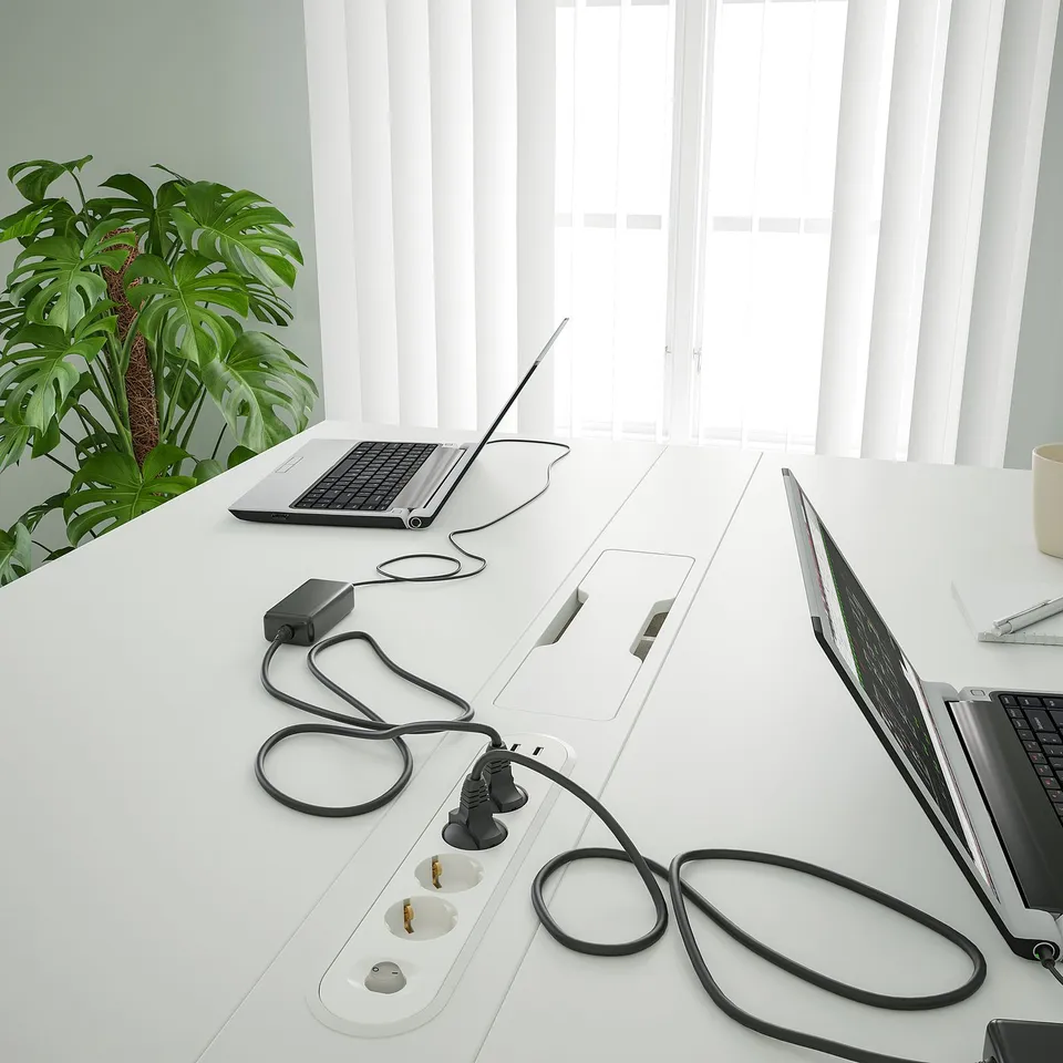 Modern office desk with power outlets and cable management, laptops, plant, and mug.