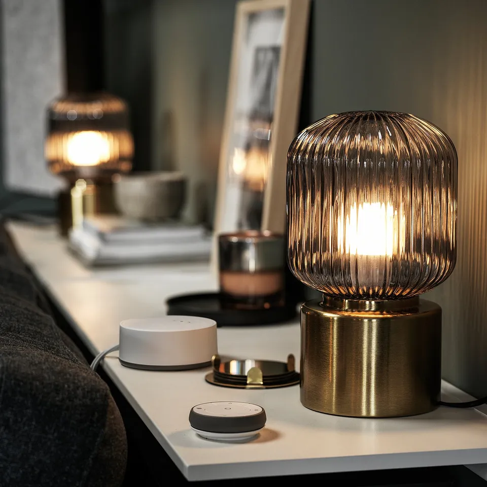 Modern interior with gold-colored SOLKLINT lamps on a bedside table, emitting soft light. A small white speaker and books are also present.