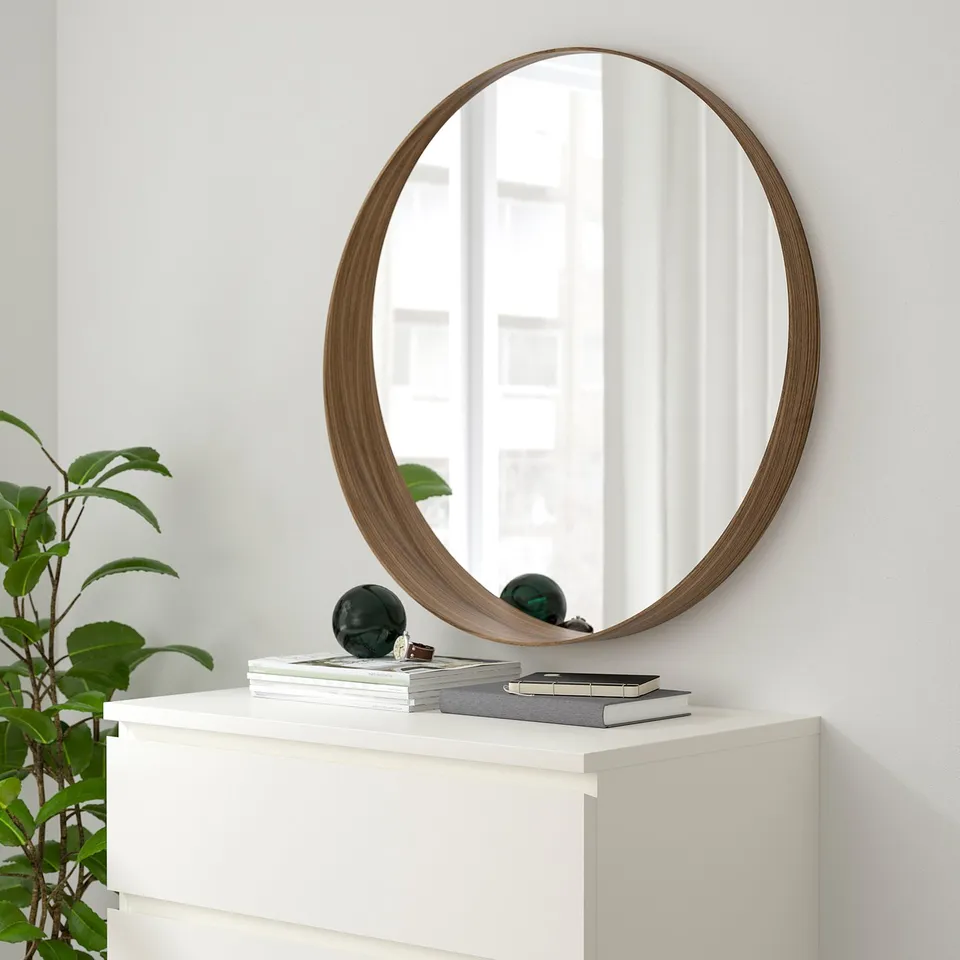 Walnut-framed STOCKHOLM mirror above white dresser, reflecting light, with decorative shelf.