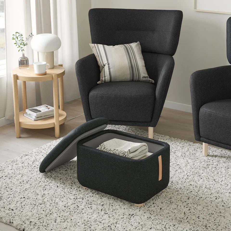 Dark grey fabric footstool with storage, wooden legs, next to a black upholstered chair with a striped pillow.