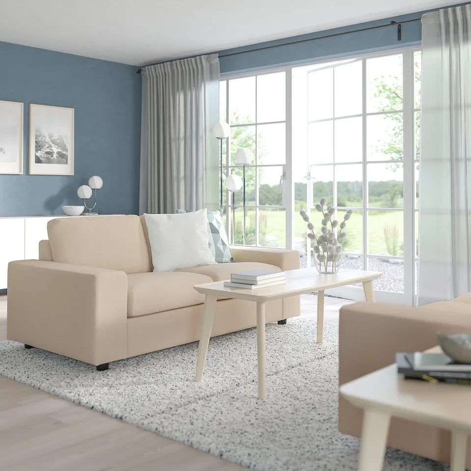 Modern living room with VIMLE beige sofa, light wood coffee table, two side tables, rug, lamp, vase, and curtains. Sofa near large windows.