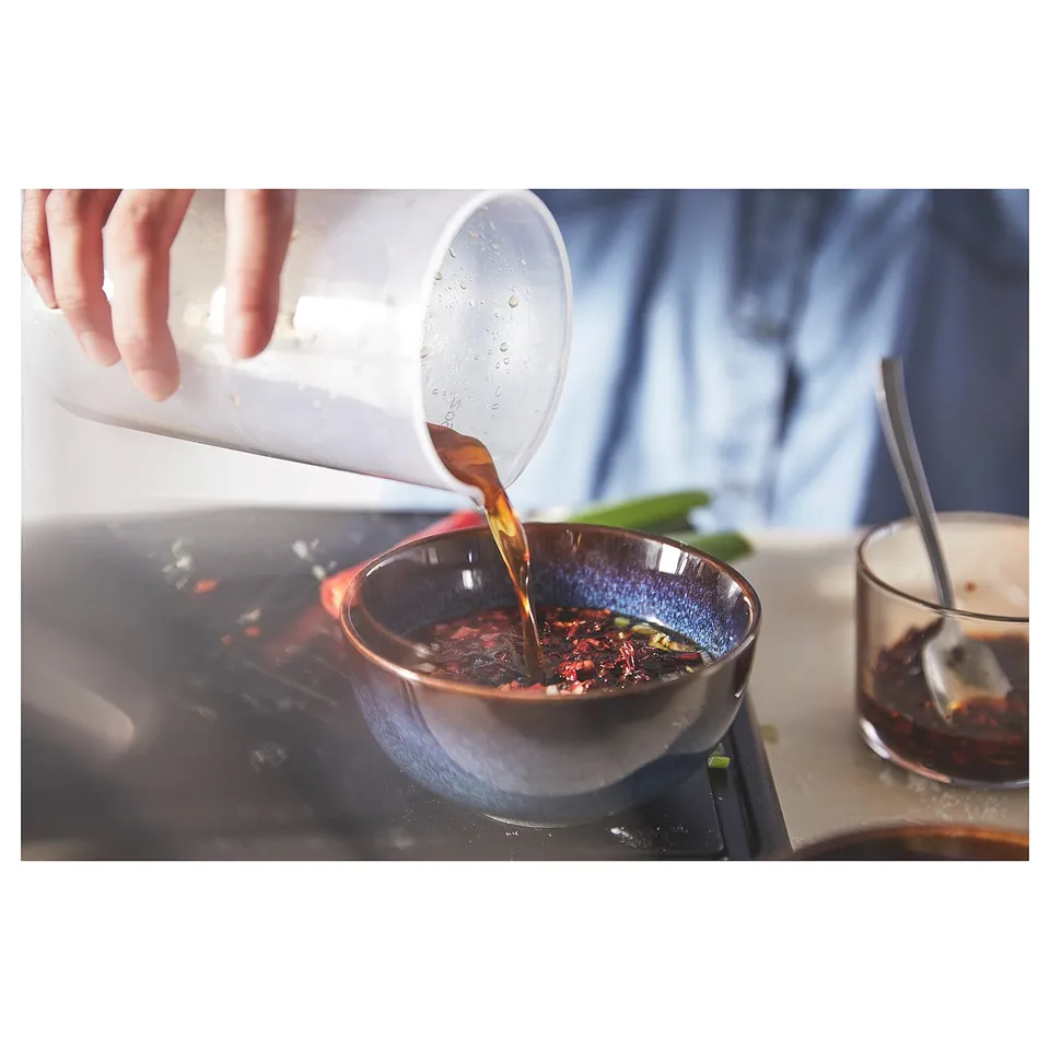 Hand pours liquid from a clear plastic pitcher into a blue glass bowl on a stove. Liquid is amber coloured.