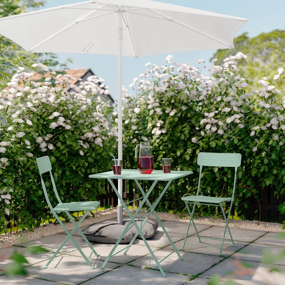 Outdoor seating set with mint green metal table and chairs, white umbrella, flowers, and drinks.