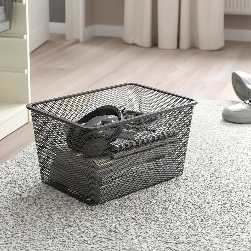 Mesh storage basket on carpet, containing headphones, near a wooden floor and white curtains.