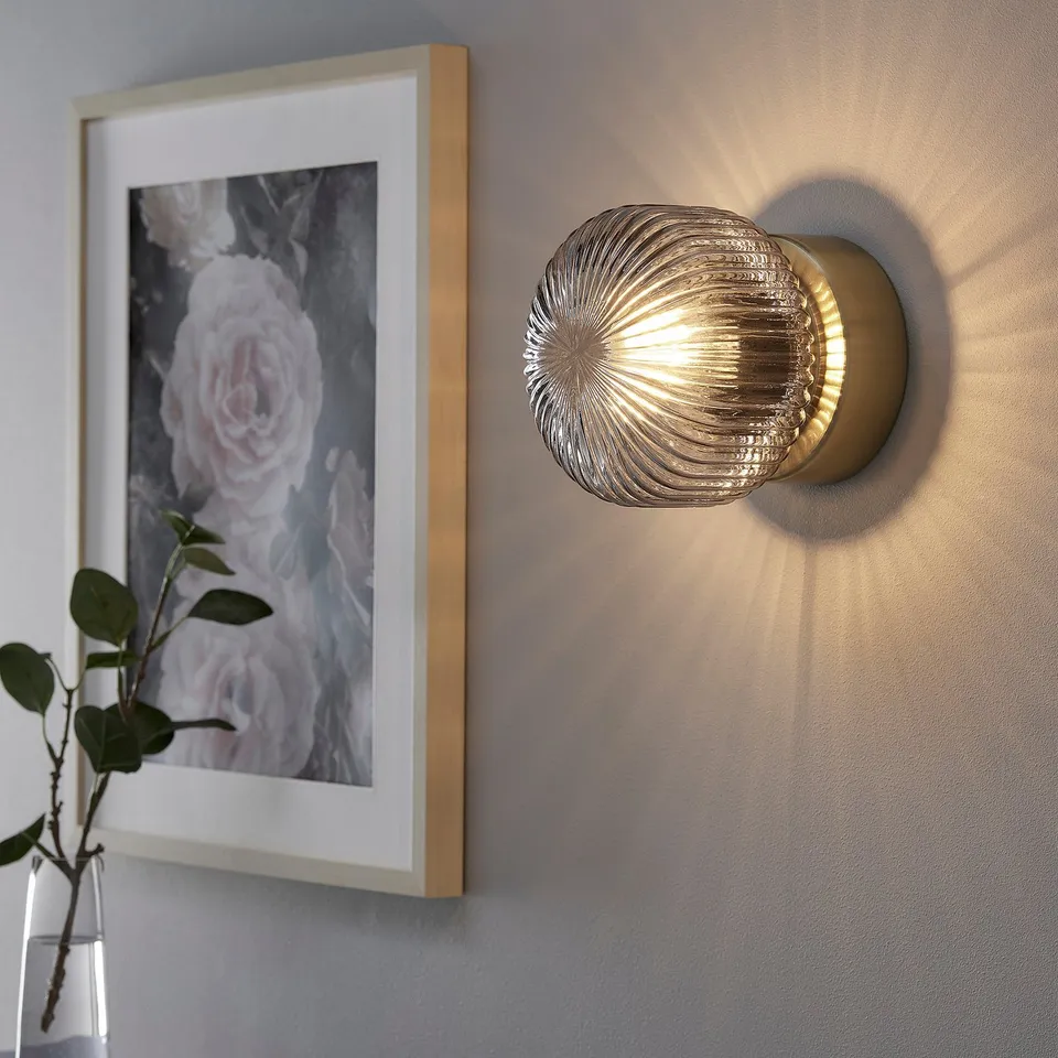 Wall-mounted brass light fixture with clear glass shade emits soft, warm light. Light bulb not included.