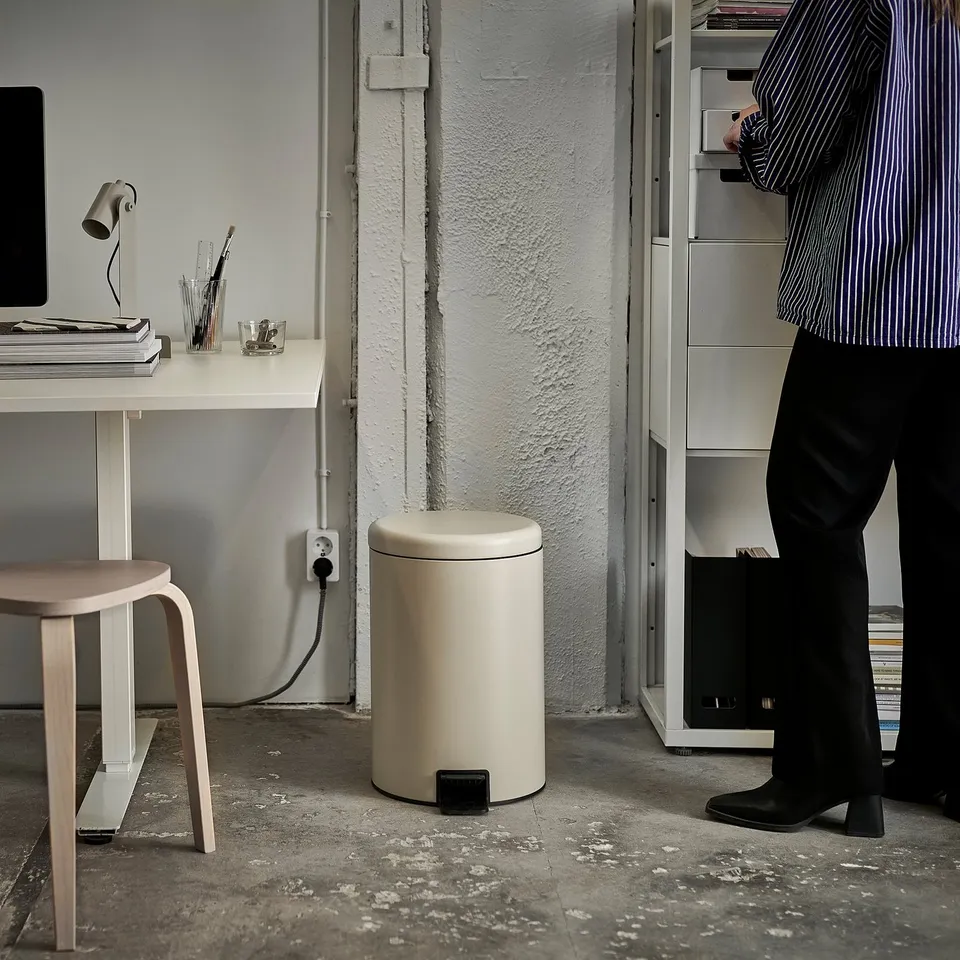 Person standing near white rubbish can with pedal, a desk with a computer and stool nearby.