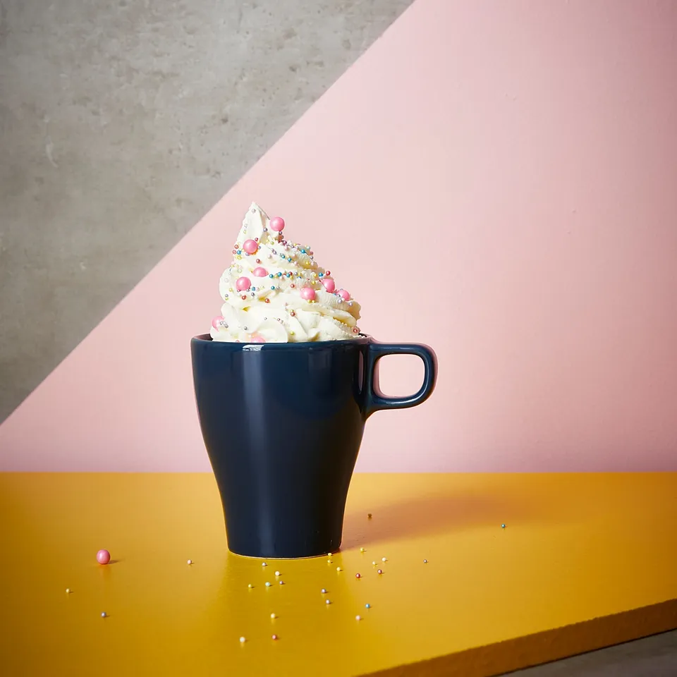 Dark blue mug with whipped cream and colourful sprinkles on a yellow table against a pink wall.