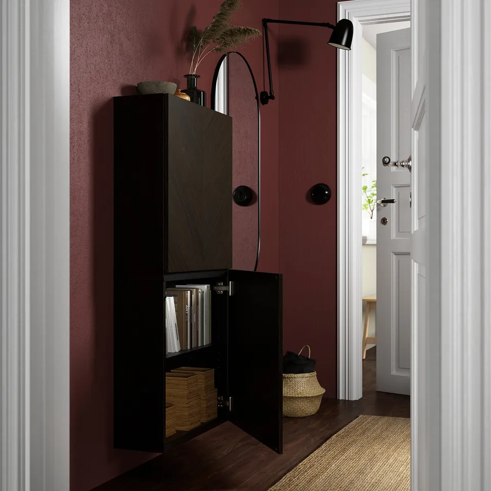 Black wall-mounted cabinet with doors open, displaying stacked books and woven baskets. Mounted on red wall, with wall lamp above.