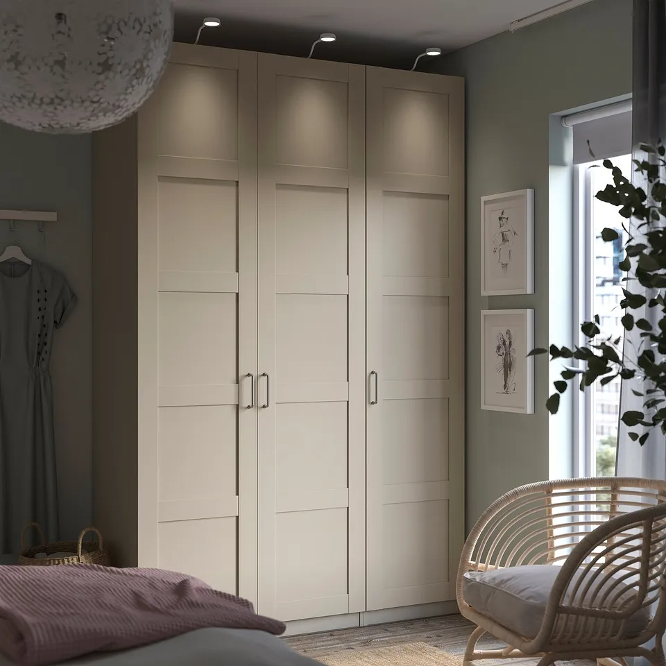 Modern bedroom with beige BERGSBO wardrobe, minimalist handles, hanging clothes, artwork, and wicker chair by window.