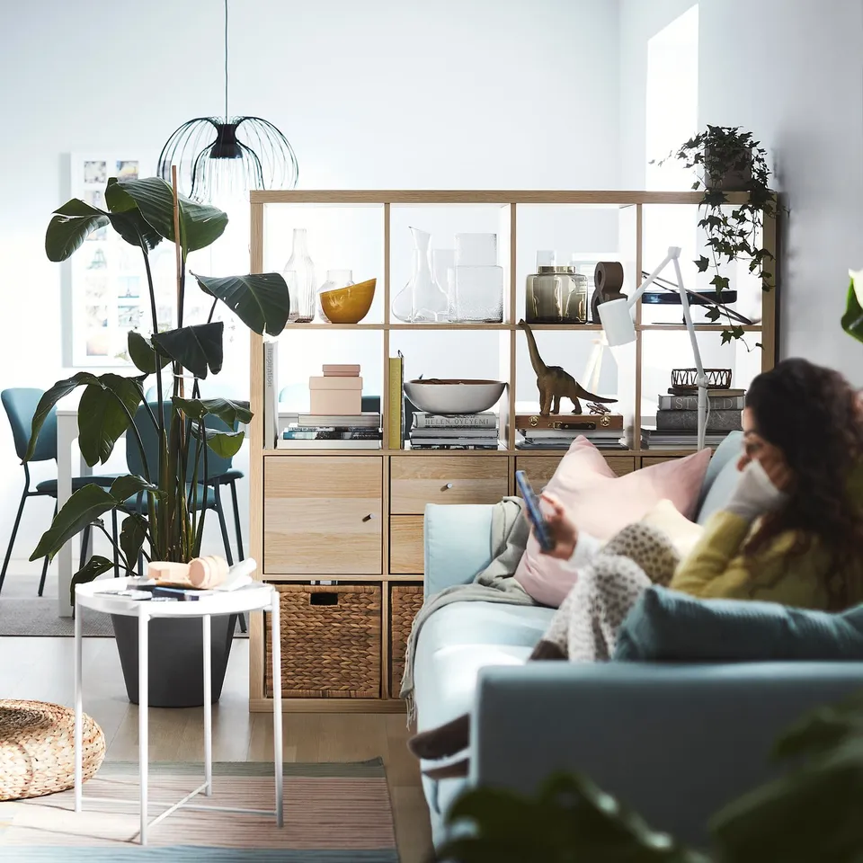 A modern living room with a KALLAX light wood shelving unit, blue sofa, and a person reading. Plants and decorative items are displayed.