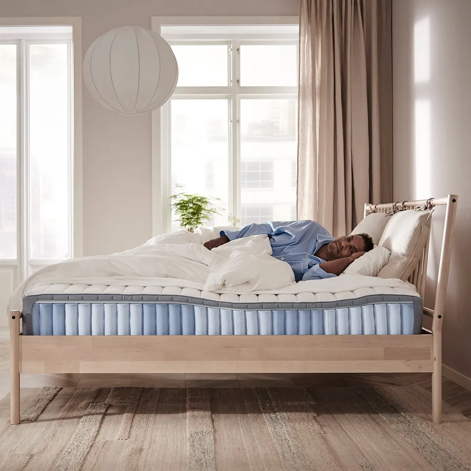 A person sleeps on a light blue VALEVÅG mattress on a wooden bed frame with white pillows and a hanging lamp.