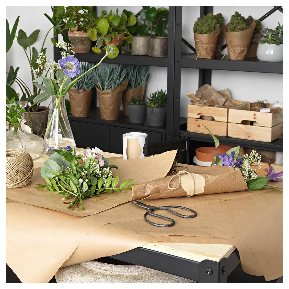 Black metal plant shelf with various potted plants and paper-wrapped flowers on table, scissors, vases, and wooden crate.