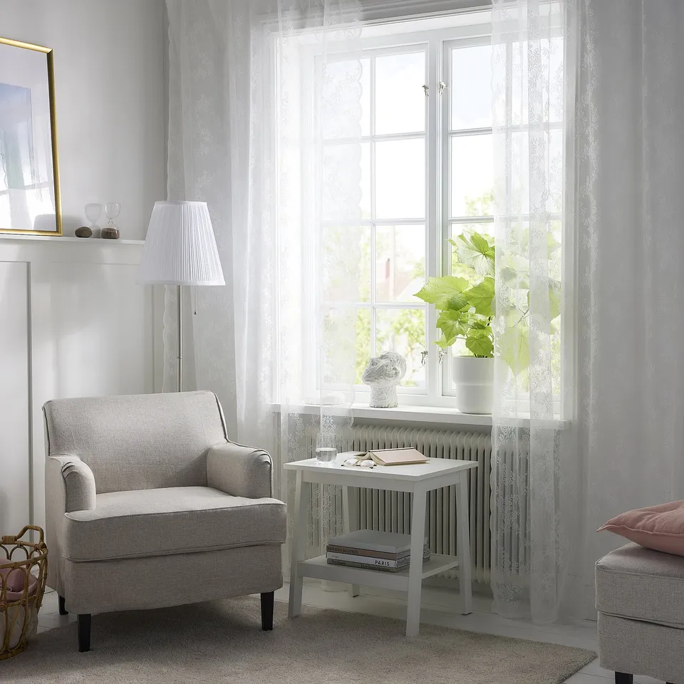 Well-lit room with grey floral curtains, armchair, books, plant, lamp.