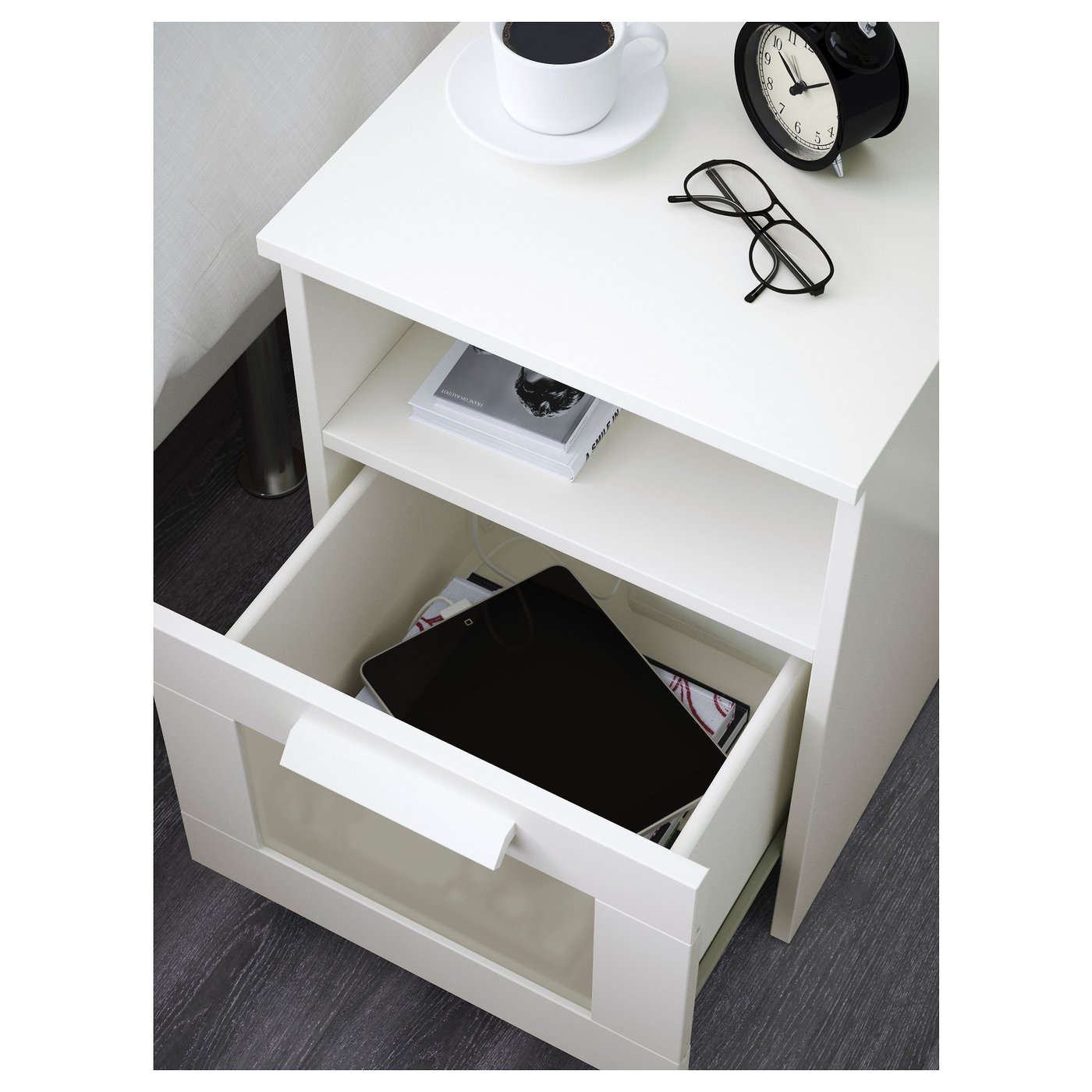 White bedside table with open drawer revealing book and tablet. Top holds coffee cup, saucer, clock, and glasses.