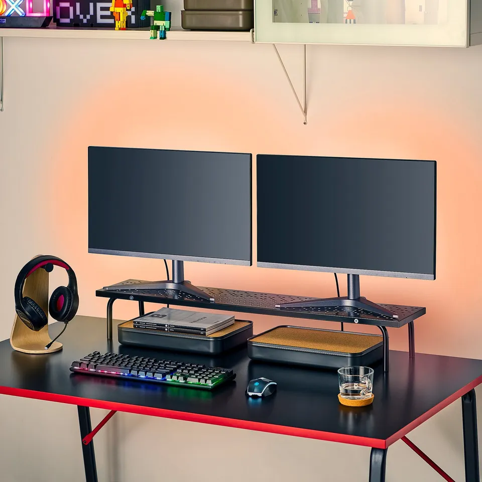 Two monitors on a stand raising them to eye level for better posture, a keyboard, mouse, headphones, and a glass on a black desk.