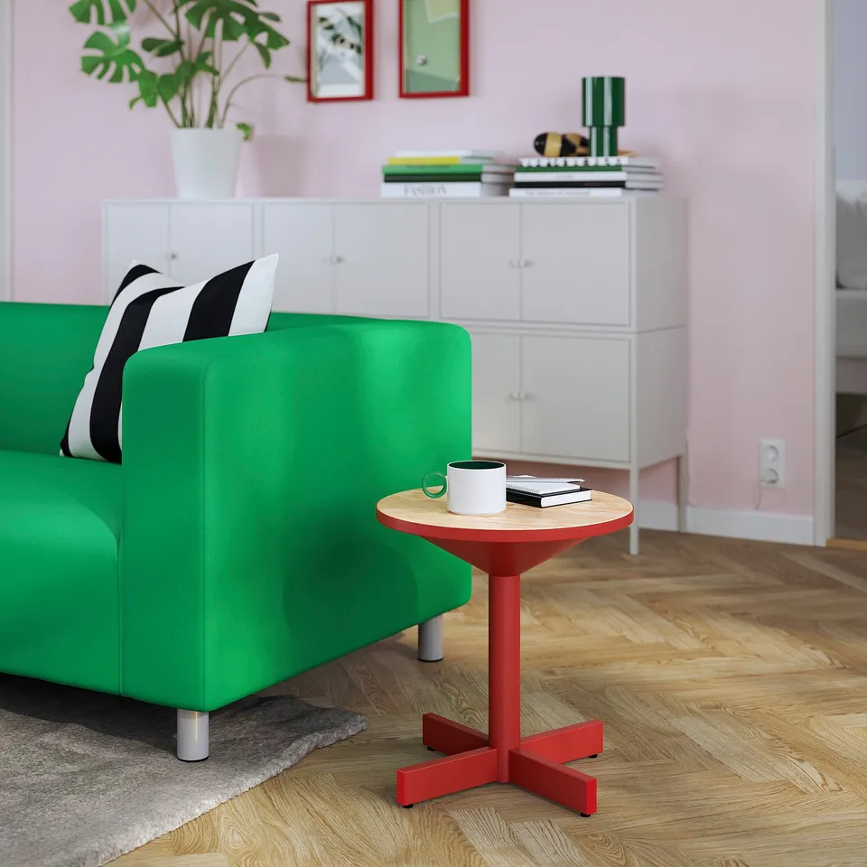 Small red side table with wooden top beside green sofa, holding coffee cup and books, in bright living space.