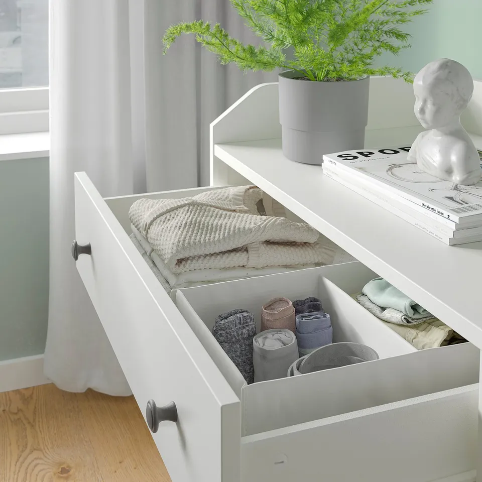 White HAUGA chest, neatly folded towels, roll-up socks, grey handles, wood floor, potted plant.