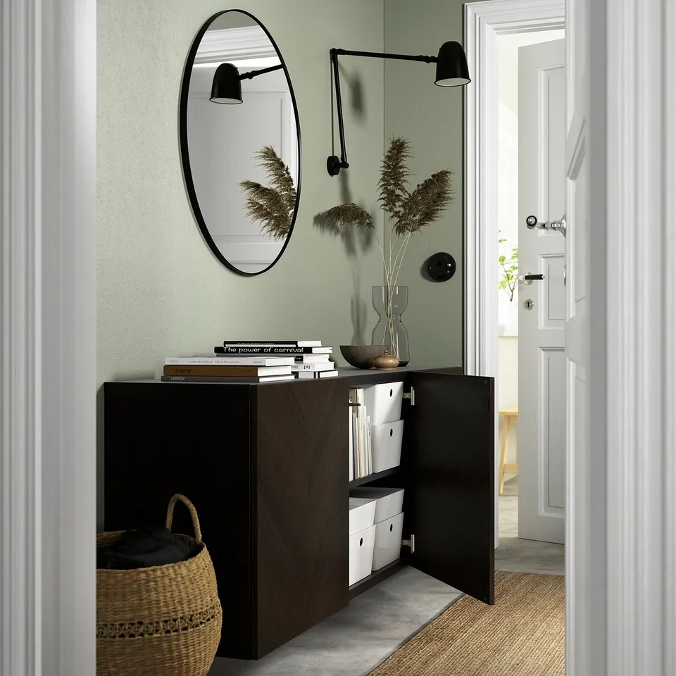 A modern hallway a wall-mounted BESTÅ storage unit with open doors revealing storage boxes. Above it, a round mirror and wall lamps enhance the decor.