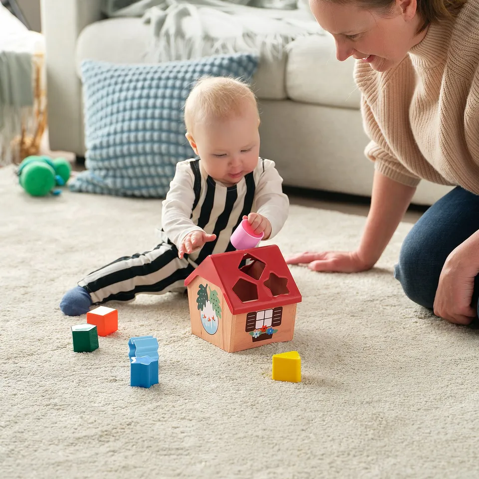 Baby plays with shape sorter toy, placing block into hole while mum watches. Toy house with colourful blocks helps learn shapes and colours.