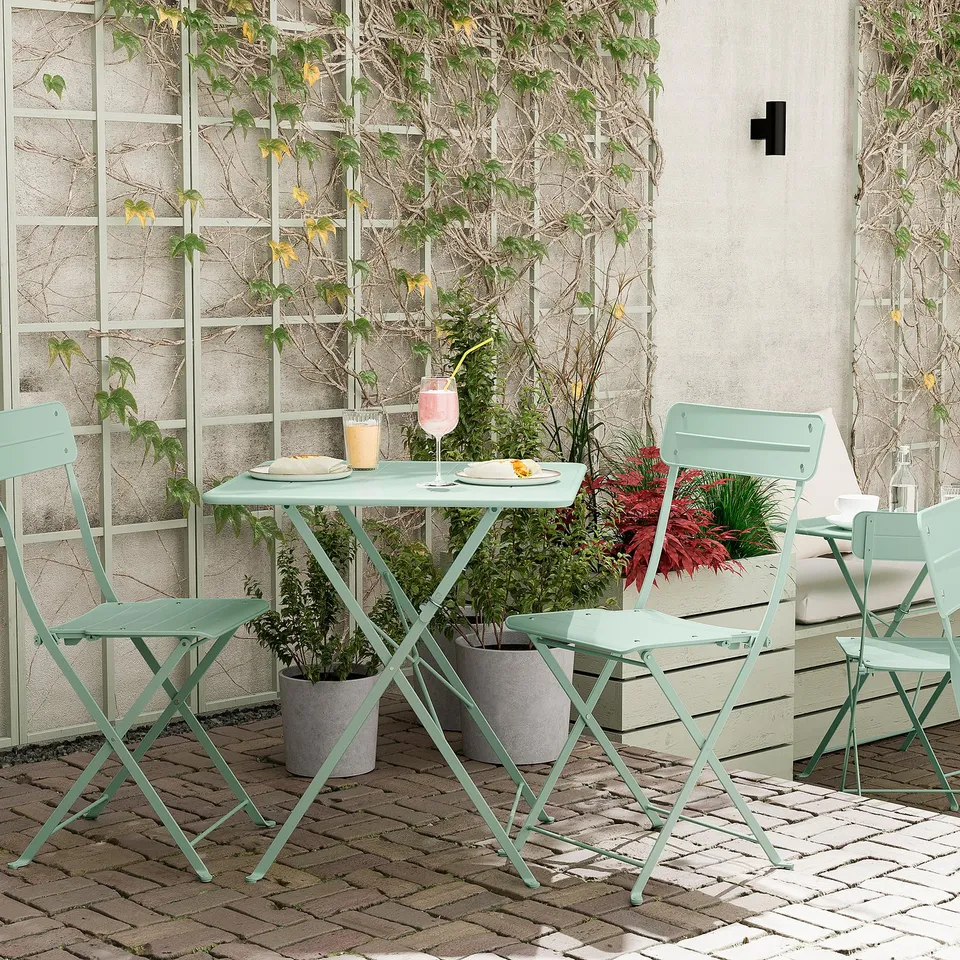 Outdoor patio with SUNDSÖ table and chairs in light green. Foldable, made of steel and polyester. Surrounded by potted plants, set on brick pavement.