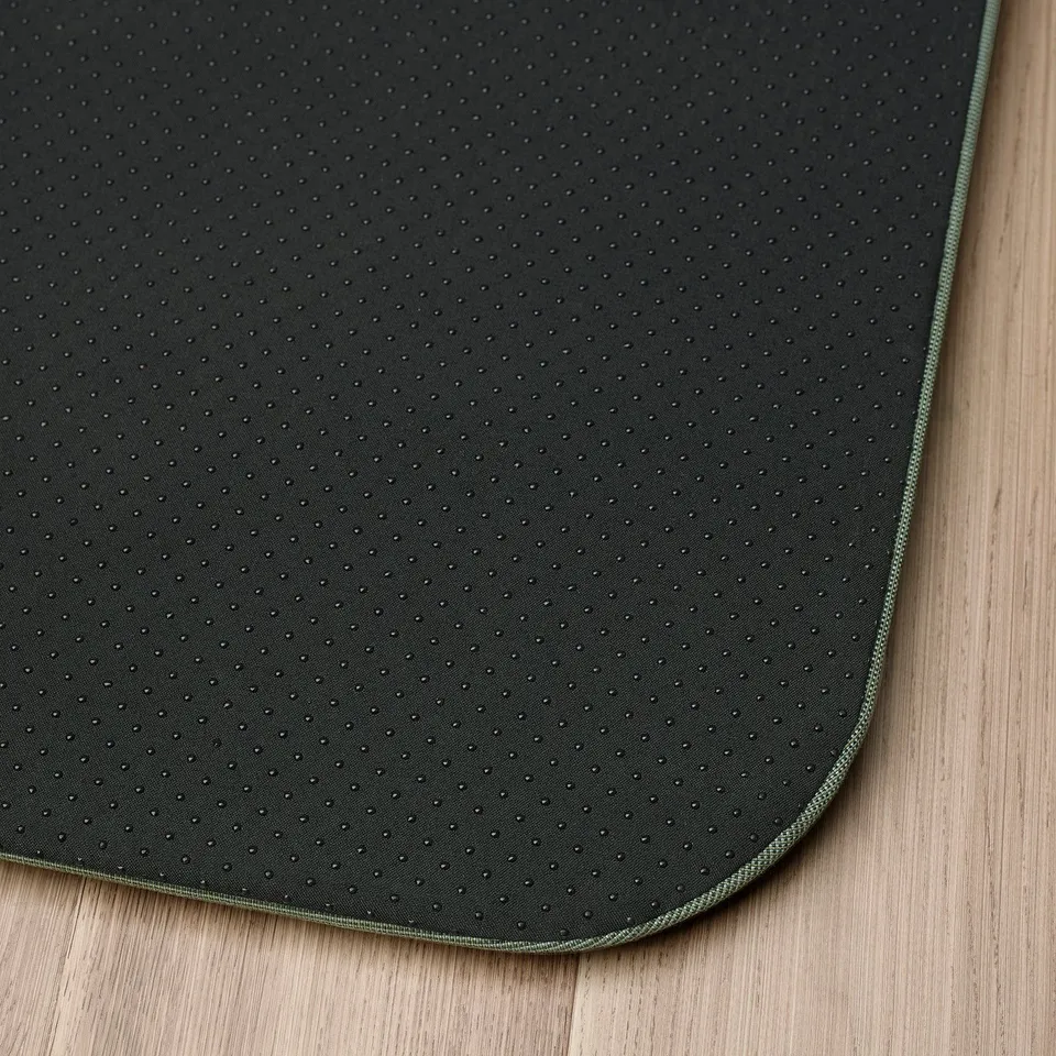 Dark anti-fatigue mat with perforated surface and green trim, placed on wood floor.