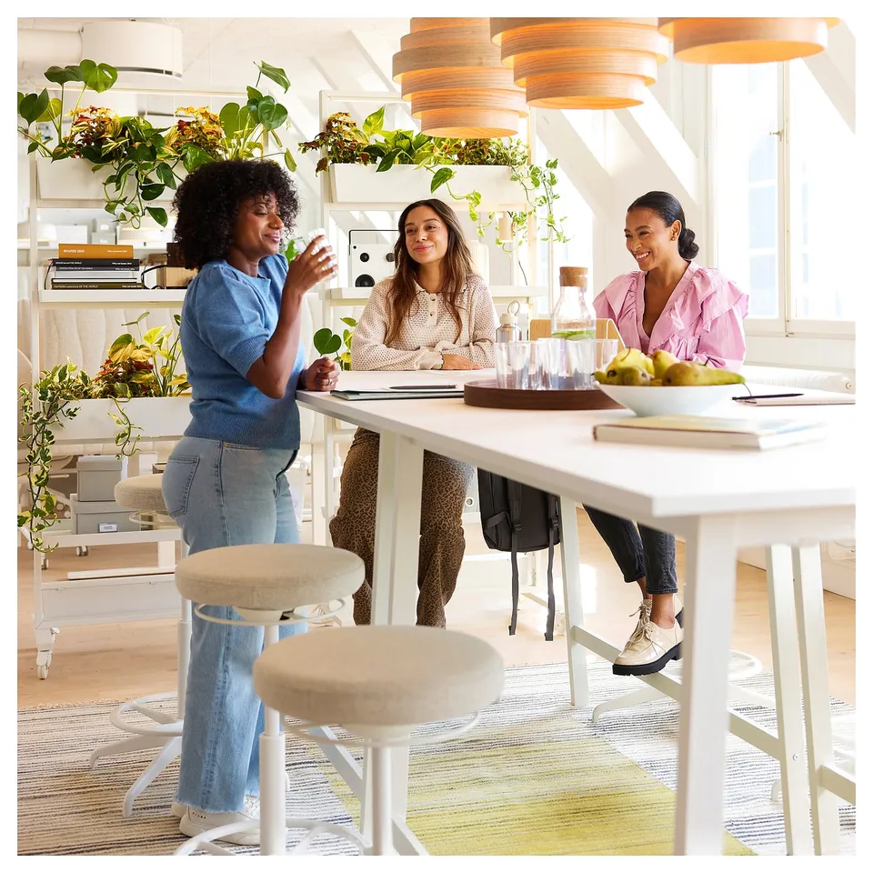 Three people chat at a sleek MITTZON high table, two sitting, one standing. Bright, airy room with plants.