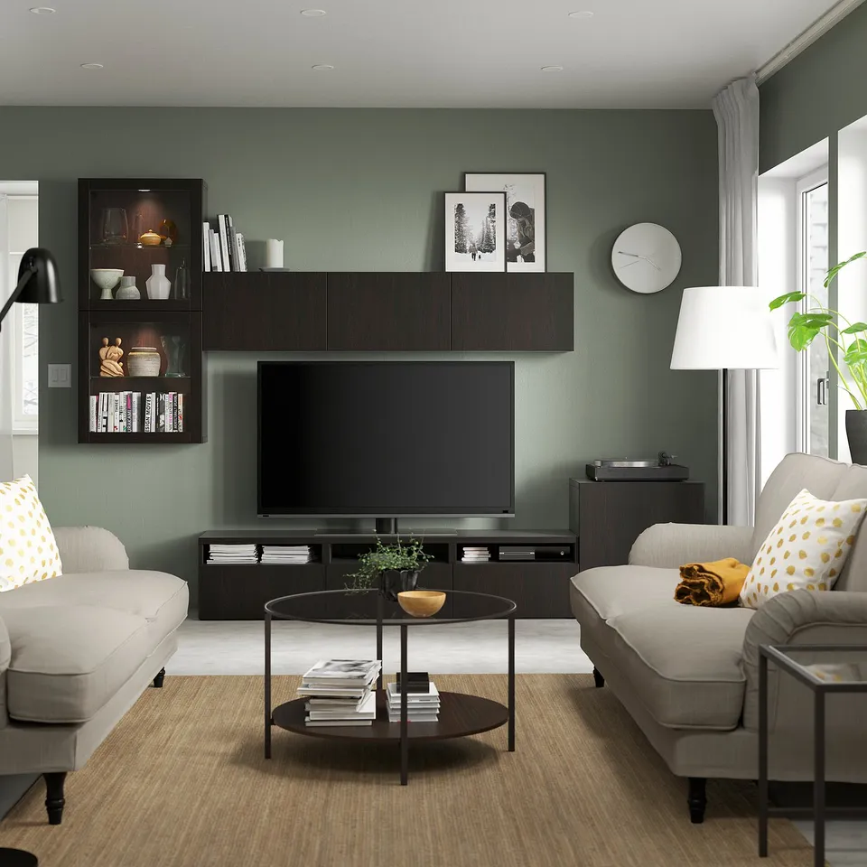 Modern living room BESTÅ tv unit with dark wood finish, flat screen tv, grey sofas, round coffee table, and green walls with large windows.