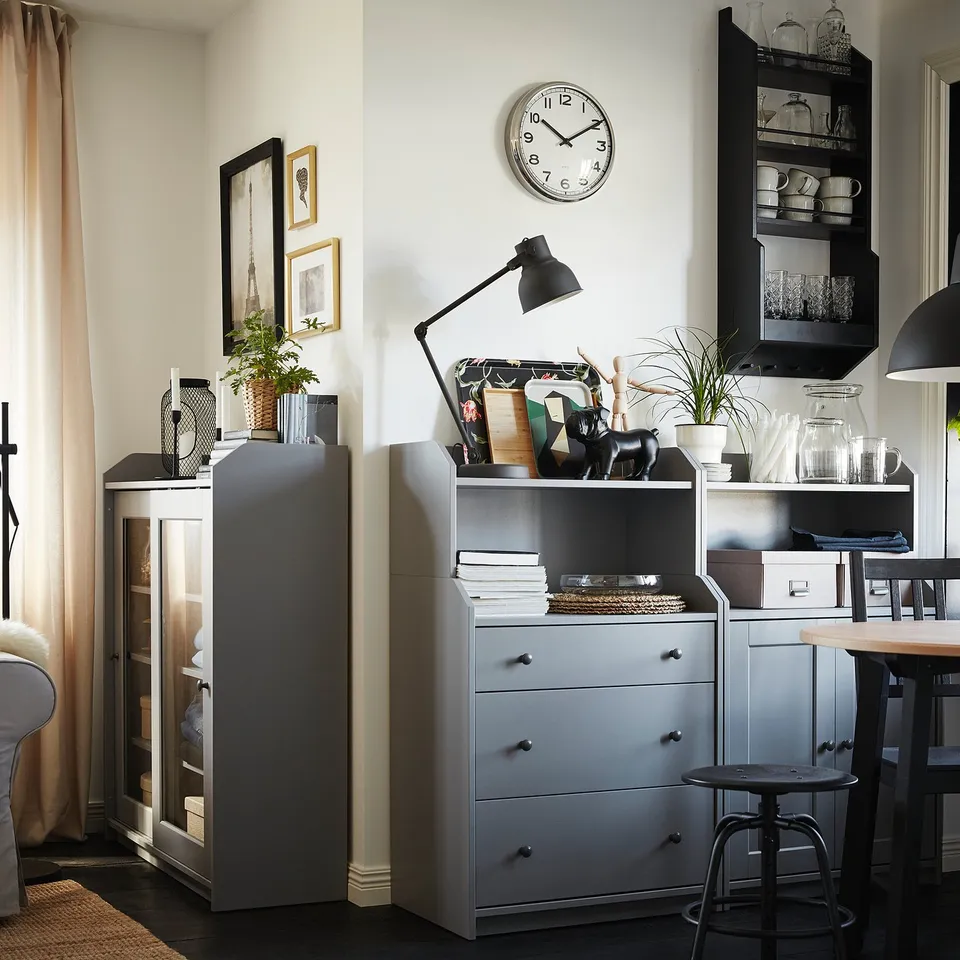 Modern living space with grey cabinets, a round wall clock, decorative items, and a dining area with hanging lamps and sheer curtains.