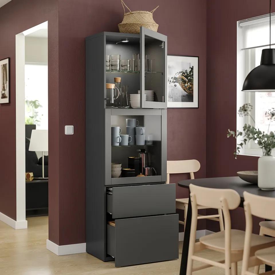 Dark wooden cabinet with glass doors displaying cups, mugs, and bowls. Two drawers beneath, set against a red wall near a dining area.