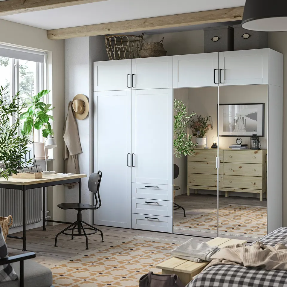Cosy bedroom with large PLATSA wardrobe. Bright white, mirrors, wood accents. Desk, chair, and bed seen, geometric rug.
