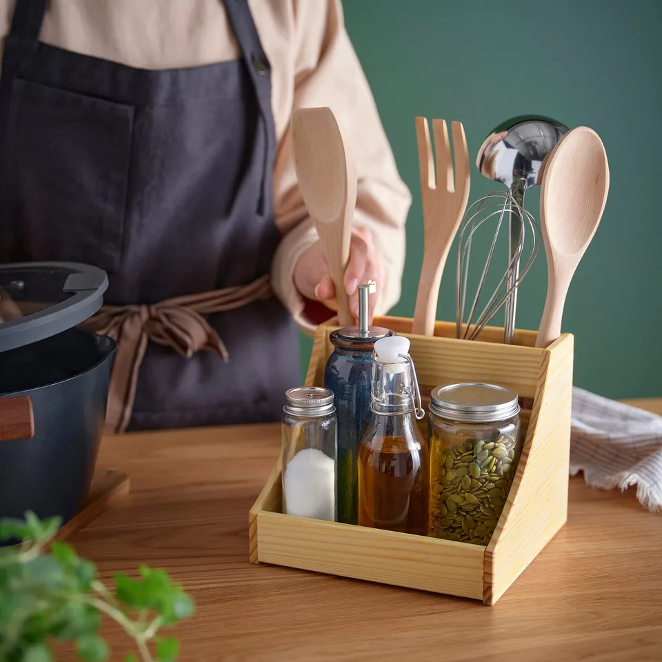 A chef in an apron holds utensils from a wooden kitchen organiser, which contains jars of spices and cooking utensils.