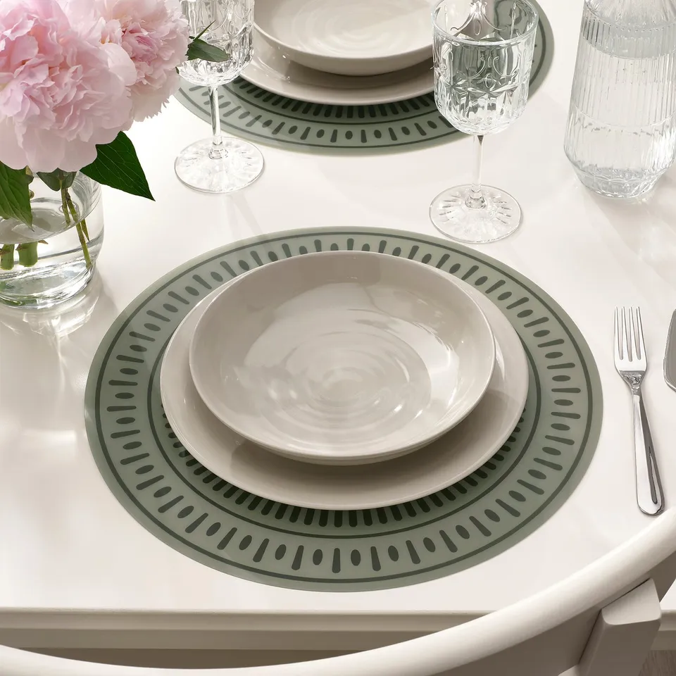 Table set with green patterned placemats, white dishes, glasses, silverware, and pink flowers.