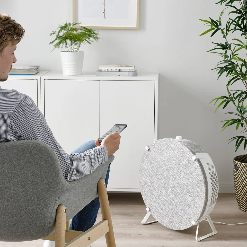 Person sits in grey chair using smartphone near white air purifier on stand.