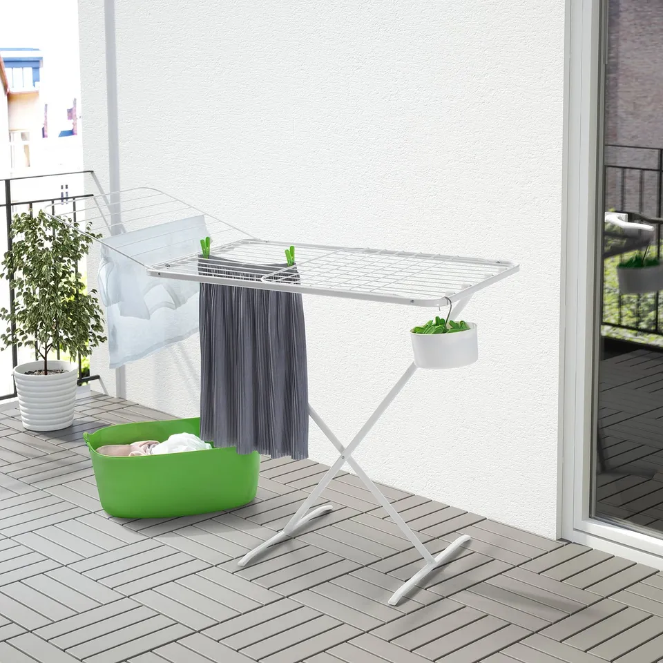 White outdoor drying rack with fold-out wings holds clothes and towels on balcony next to plant and laundry basket.