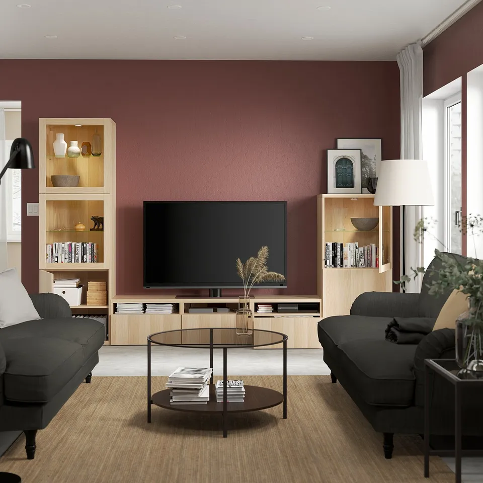 Modern living room with dark red walls, wooden shelves & drawers, grey couches, glass coffee table, and tall lamp.