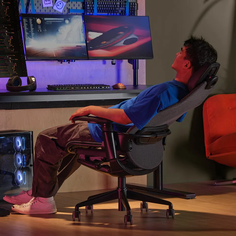 Person reclines in ergonomic gaming chair with adjustable supports, before dual computer monitors displaying games.