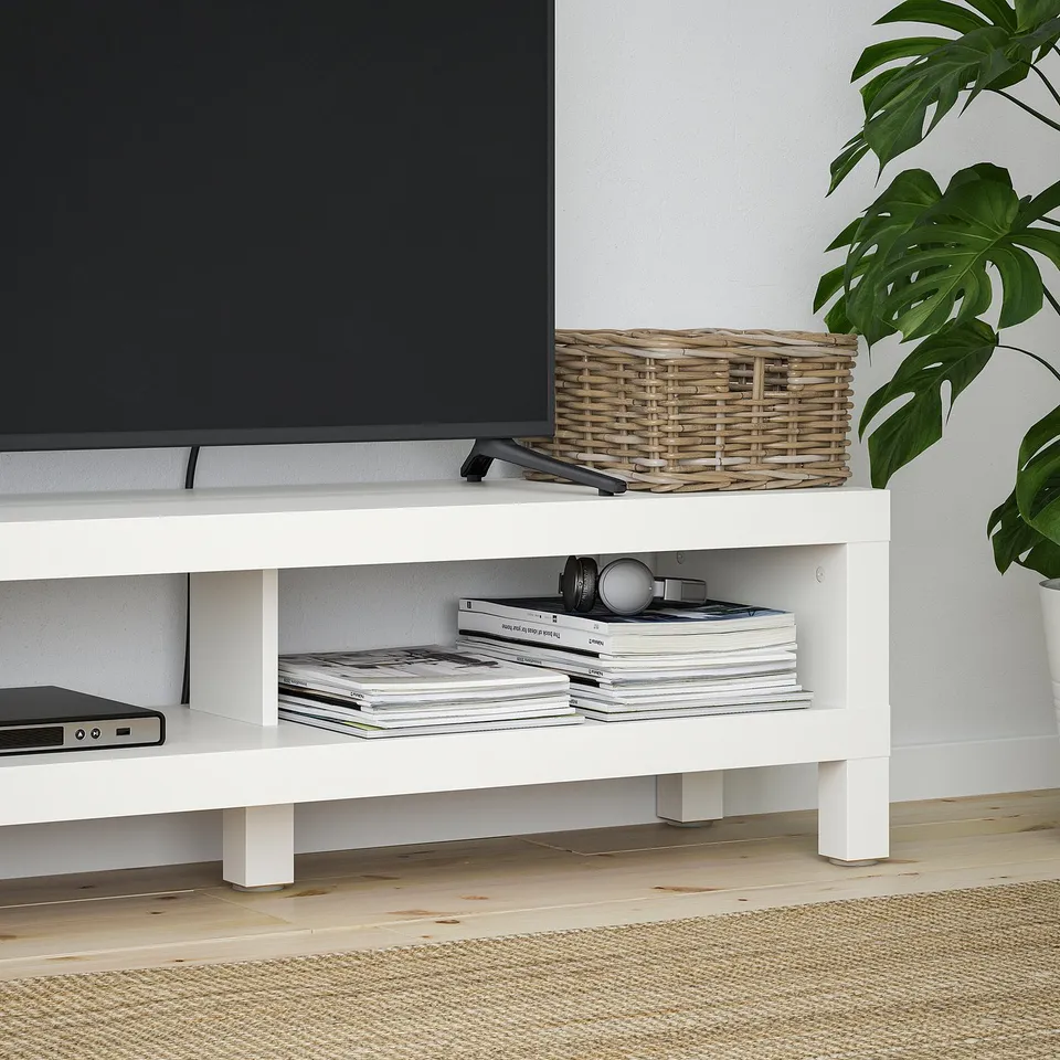 A white tv stand with open storage shelves holds a modern tv, a wicker basket, and stacks of magazines.