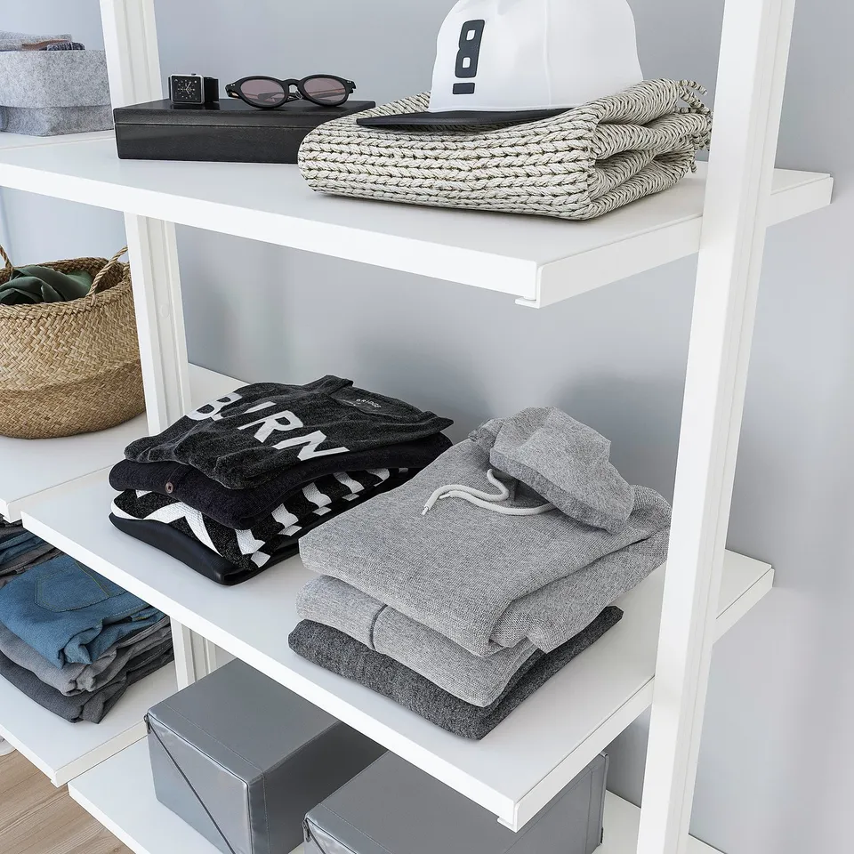 A neatly organised closet with folded sweatshirts and a baseball cap on white shelves.