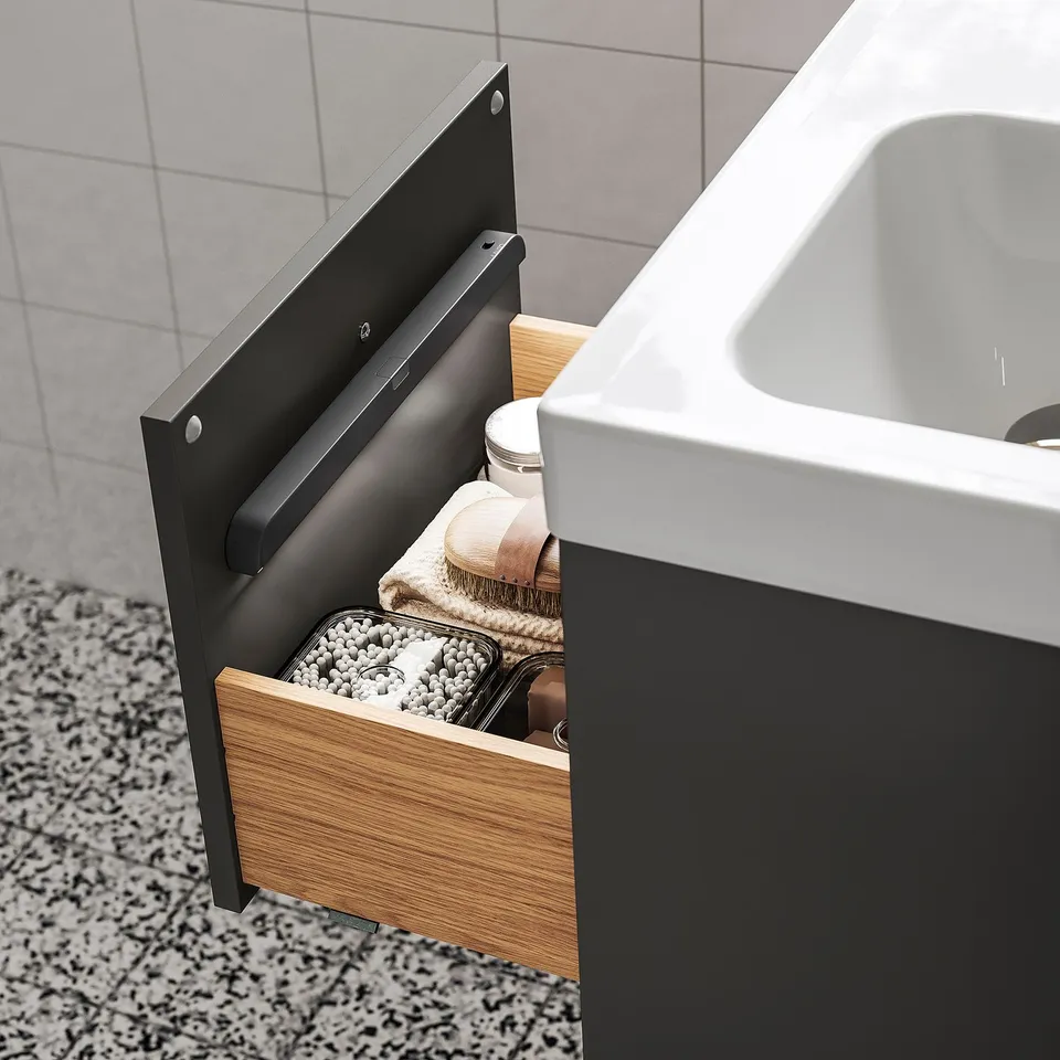 Open wooden drawer reveals organised bathroom items in dim lighting, with a modern white sink nearby.