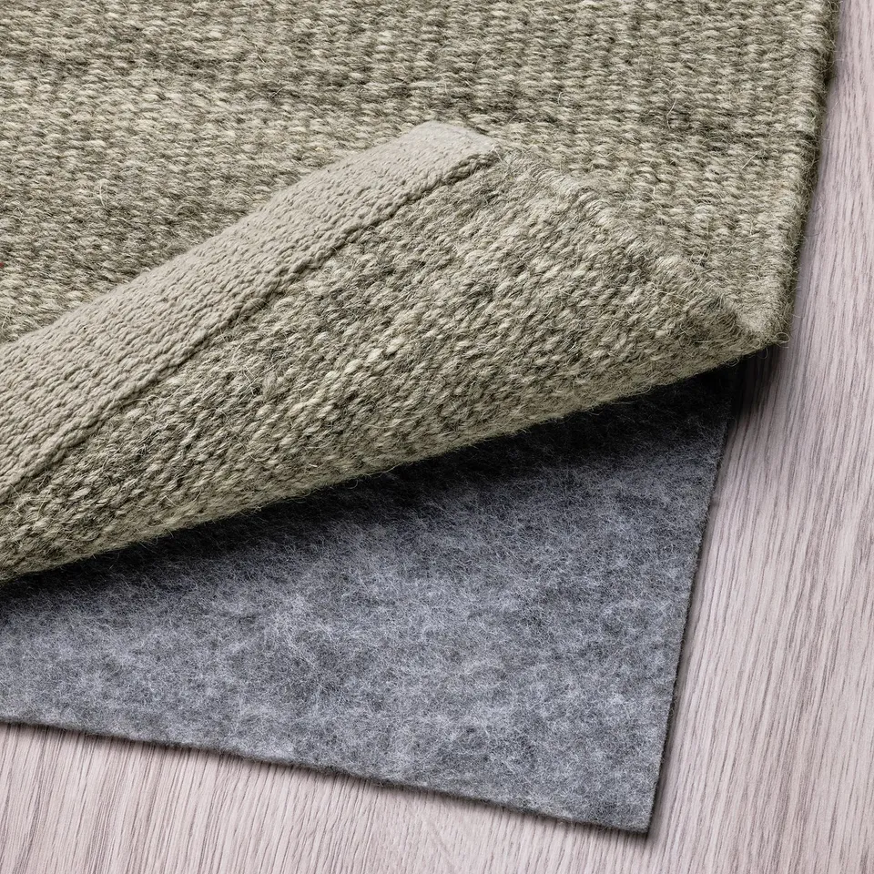 A folded grey wool rug is displayed, showcasing its underside with an anti-slip felt pad attached, ensuring stability and comfort.