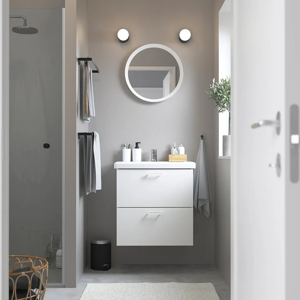Modern white ENHET bathroom vanity with two drawers, mounted on wall, smaller than shower stall, with round mirror and lights above.