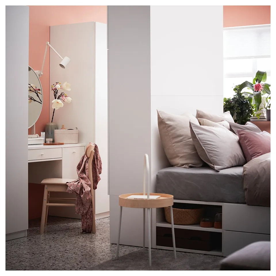 Bedroom with grey bedding, pink accents, wooden side table, white dressing table with mirror, lamp, and plant.