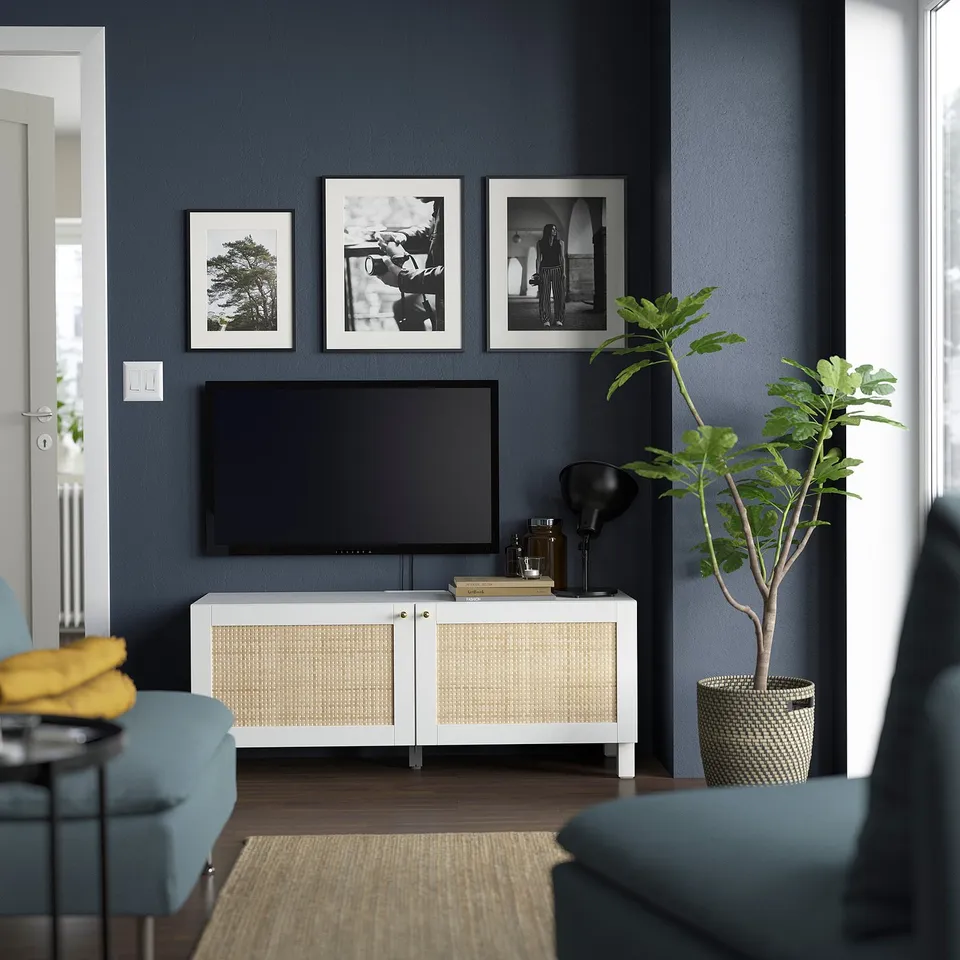 A modern living room a BESTÅ tv bench in white and cane with a flat-screen tv and framed art, complemented by blue seating.