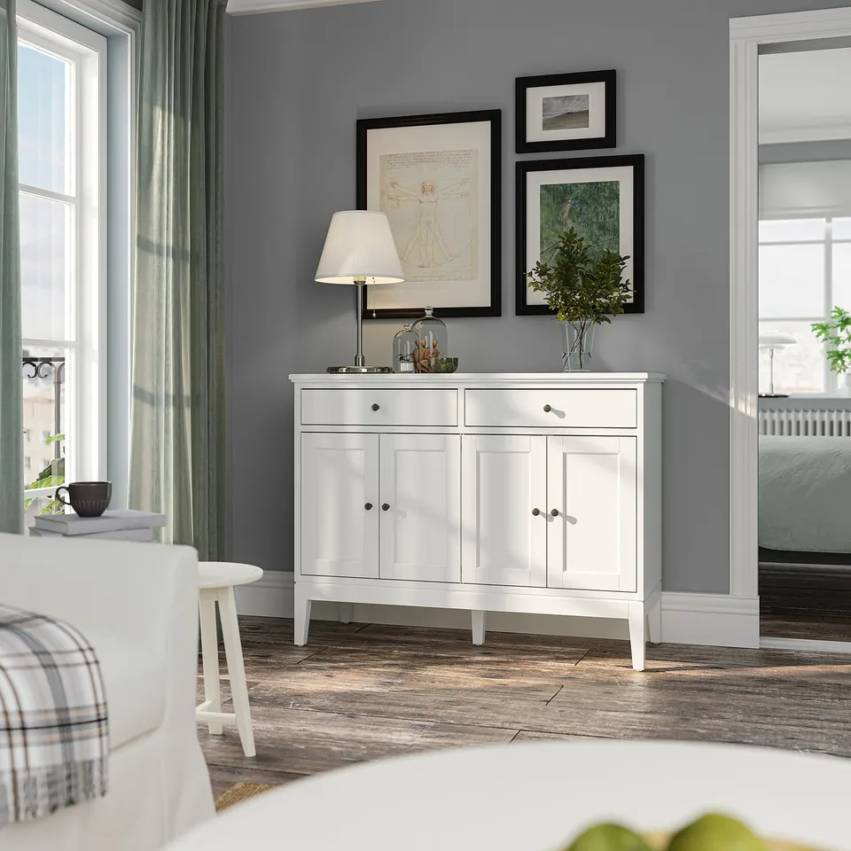Modern white sideboard with black handles, featuring framed art and lamp on top, adjacent to a dining table with a plaid chair.