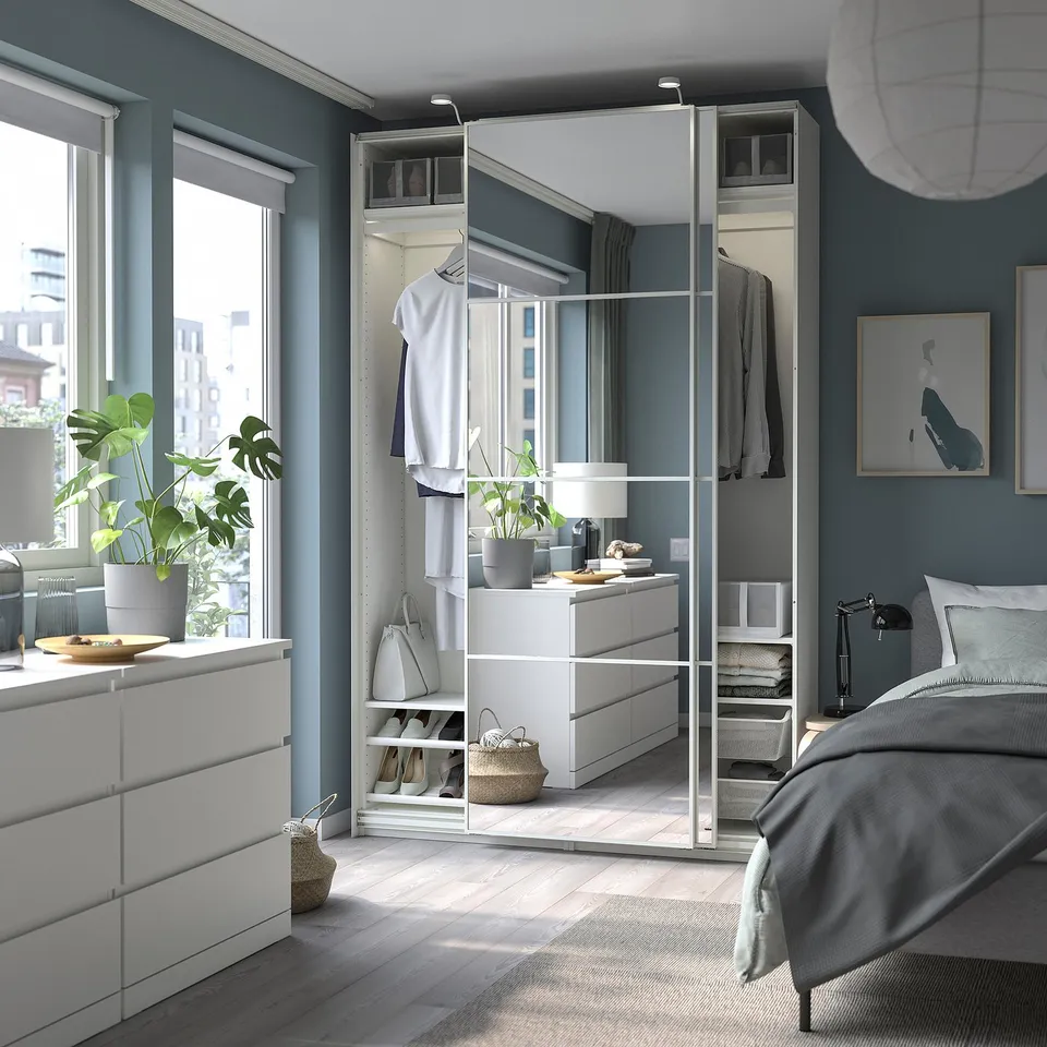Modern bedroom with grey walls, white PAX wardrobe, mirrored doors. Cosy bed, rug, large plants by window.