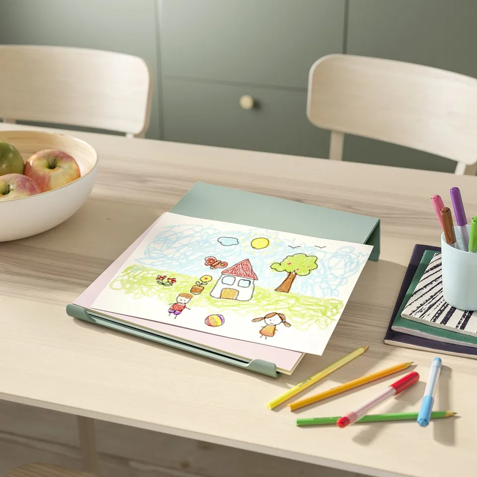 On a light-wood desk, a childs drawing, coloured pencils, and a bowl of apples sit. Two stackable chairs are placed nearby.