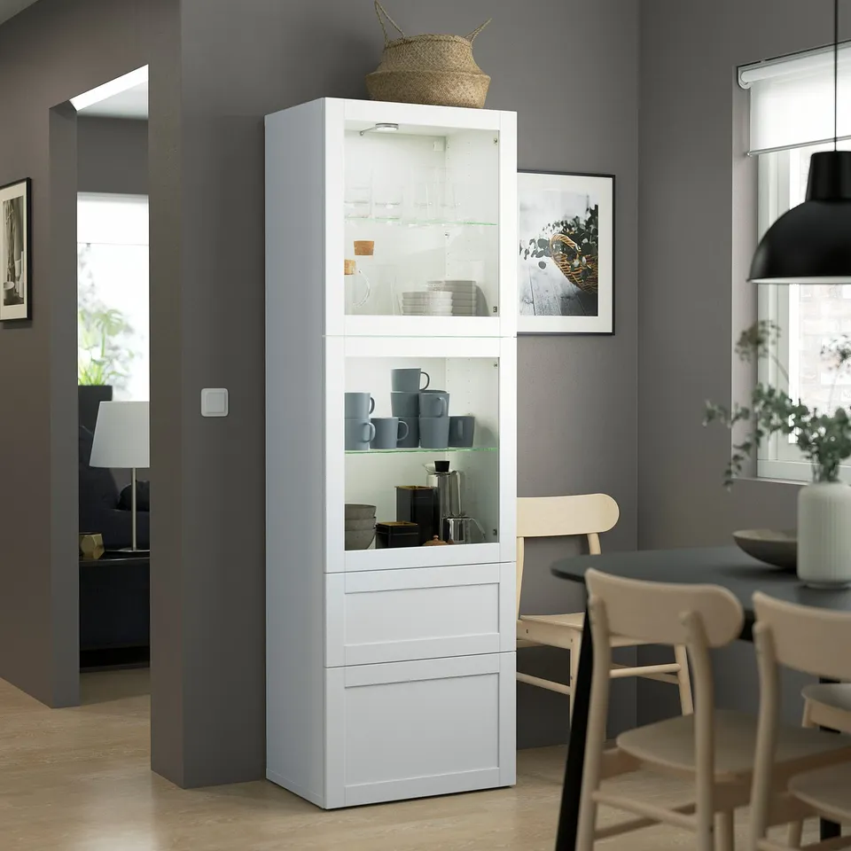 White BESTÅ cabinet, tall and narrow, with glass doors and shelves, displayed in a dining room. Surrounded by wooden chairs and a table.