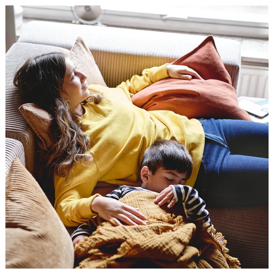 A person and child relax on a yellow JÄTTEBO modular sofa, featuring cushions and a cosy blanket.