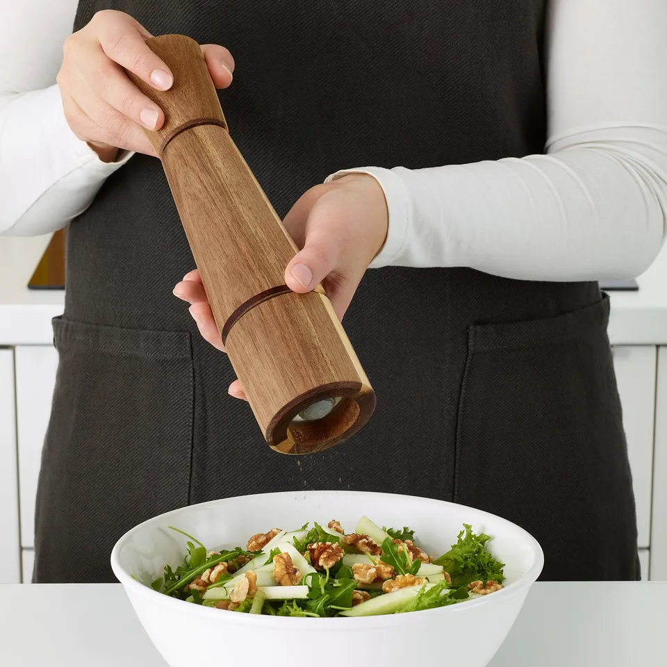 A person grinds pepper into a salad bowl using a wooden pepper mill with a ceramic grinder.