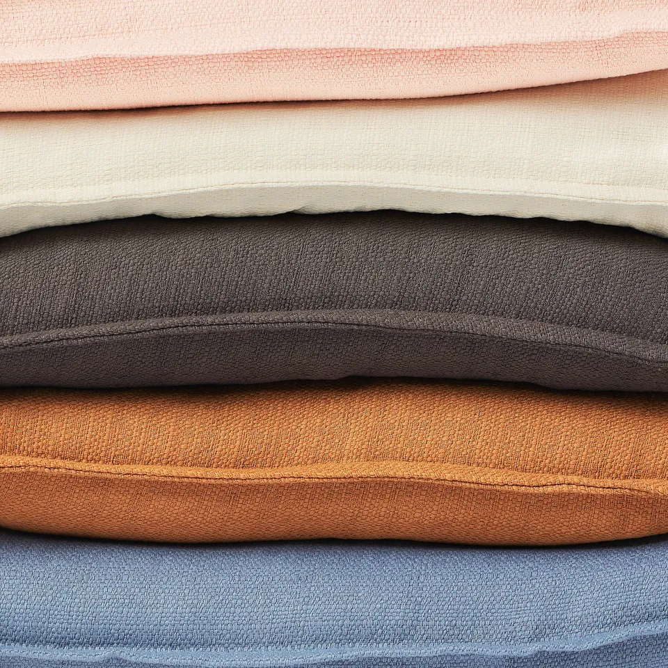 Stacked pillows in light pink, beige, grey, brown, and blue. Each has a textured surface and zip detail.